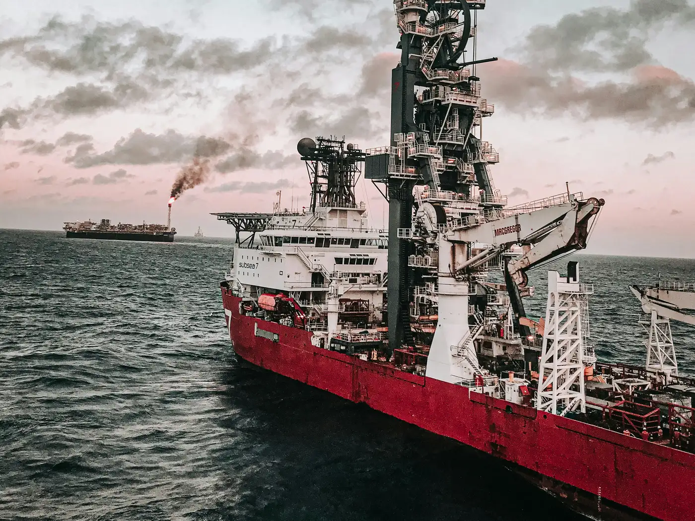 An offshore construction vessel at dusk, a production platform flaring behind it